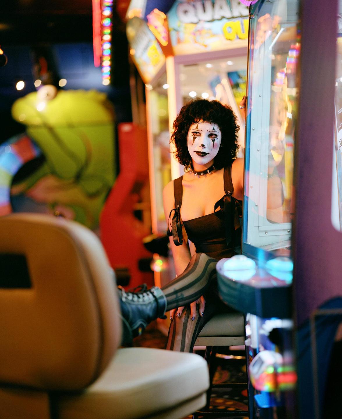 A woman in clown makeup and costume sits in an arcade next to a claw machine.