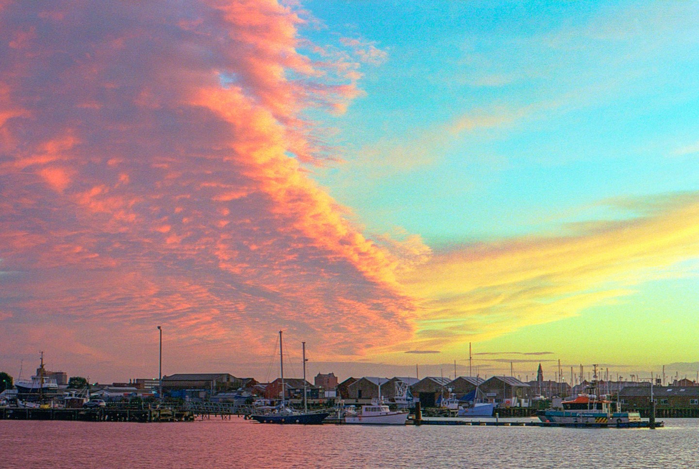 A vibrant sunset over a harbor with boats docked along the shore and buildings in the background.
