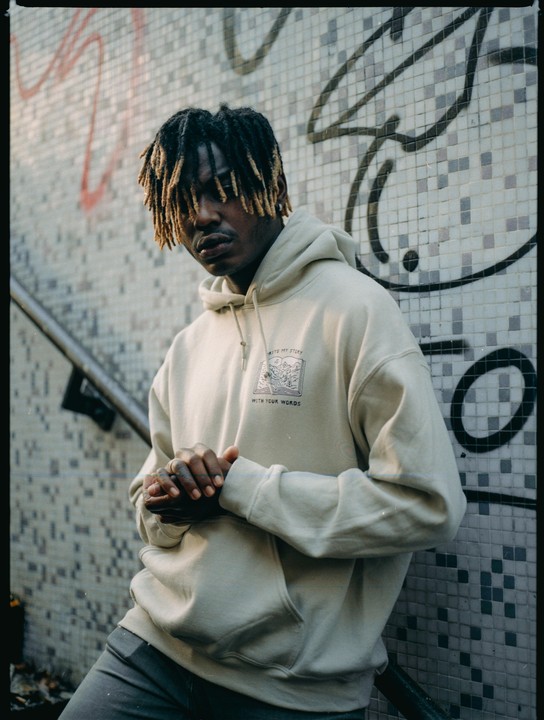 A man with dreadlocks and a light-colored hoodie leans against a tiled wall with graffiti.