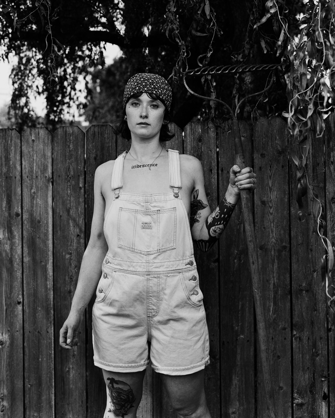 A woman in overalls stands in front of a wooden fence, holding a garden tool.