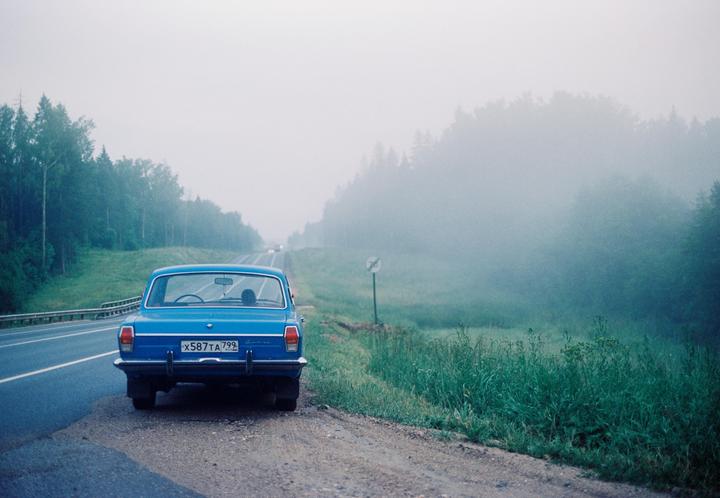 A blue car is parked on the side of a road next to a forest shrouded in fog.