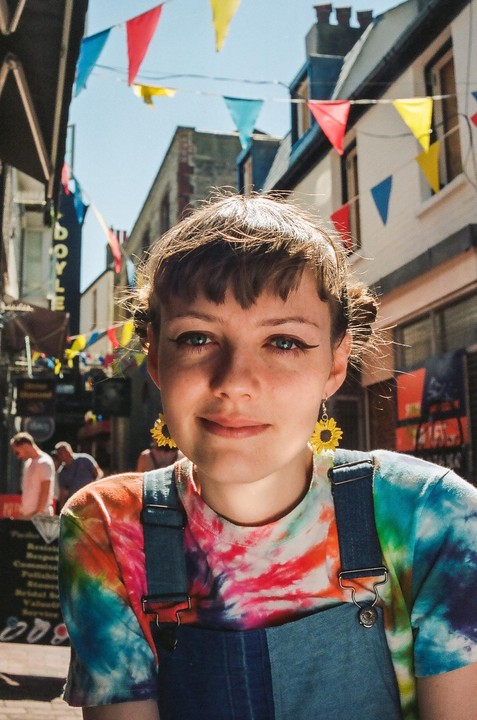 A woman with sunflower earrings and a tie-dye shirt smiles at the camera on a street decorated with colorful bunting.