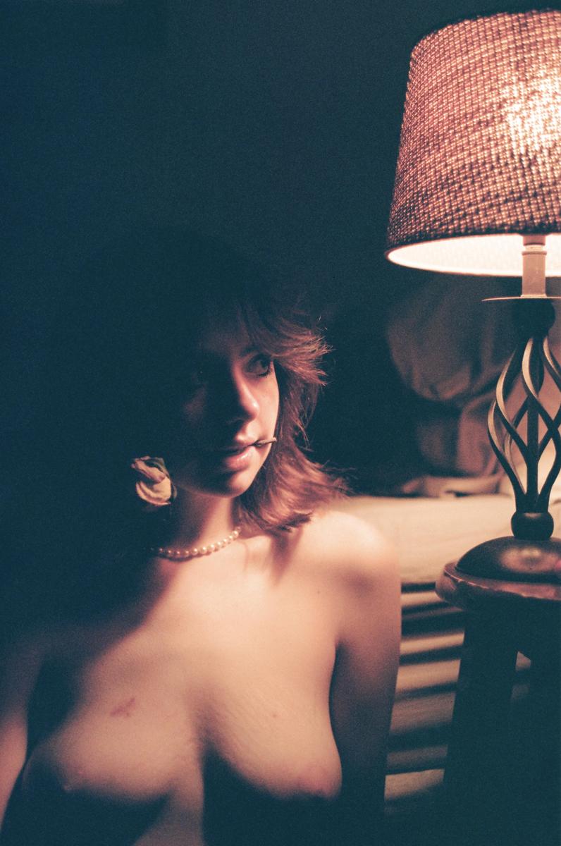 A woman with a pearl necklace and a flower in her mouth is illuminated by a lamp.