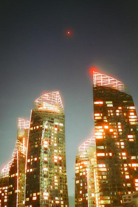 Four skyscrapers with illuminated windows and red lights on their roofs stand against a dark sky with a single red light.