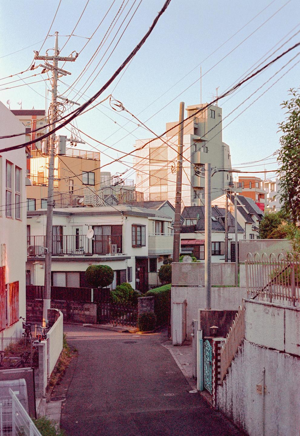 A narrow street winds through a dense urban neighborhood with numerous power lines overhead.