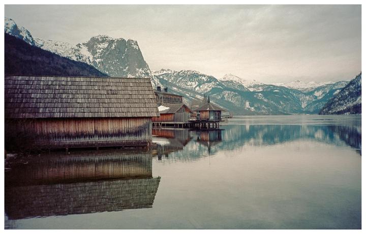 Wooden boathouses sit on a calm lake with snow-capped mountains in the background under a cloudy sky.