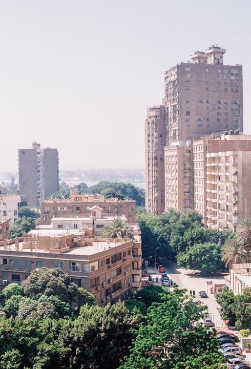 An aerial view shows a city street lined with trees and buildings, with a body of water in the distance.