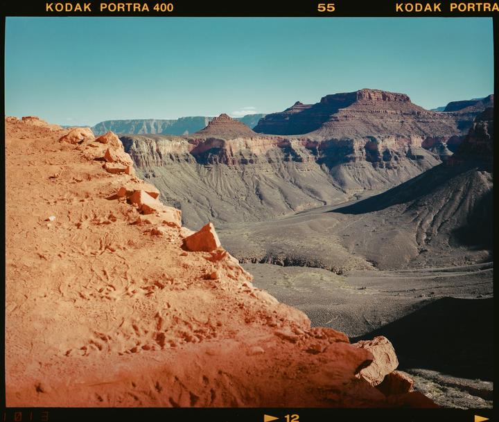 A vast canyon landscape features a red rock formation in the foreground and distant mesas under a clear sky.