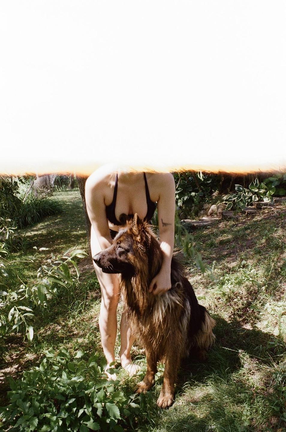A woman in a bikini top stands in a garden, holding a large dog.