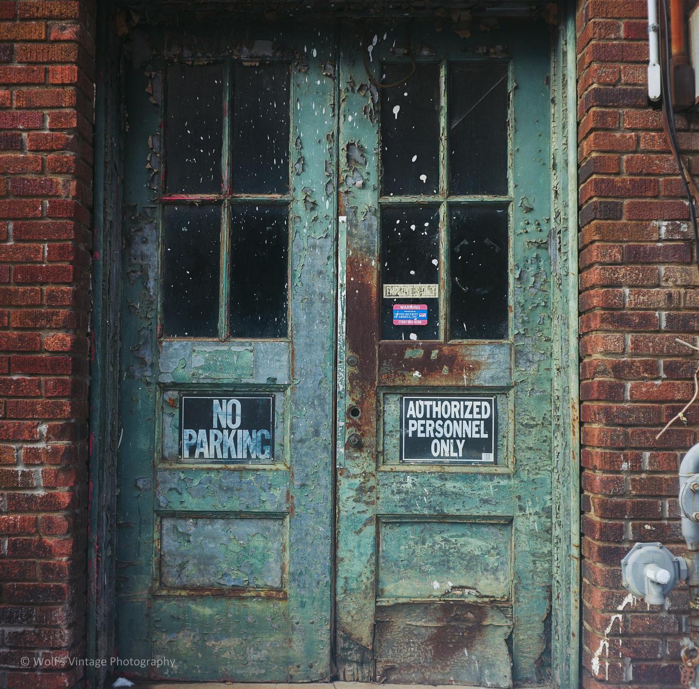 A pair of weathered green doors with peeling paint and glass panes are set into a brick wall.