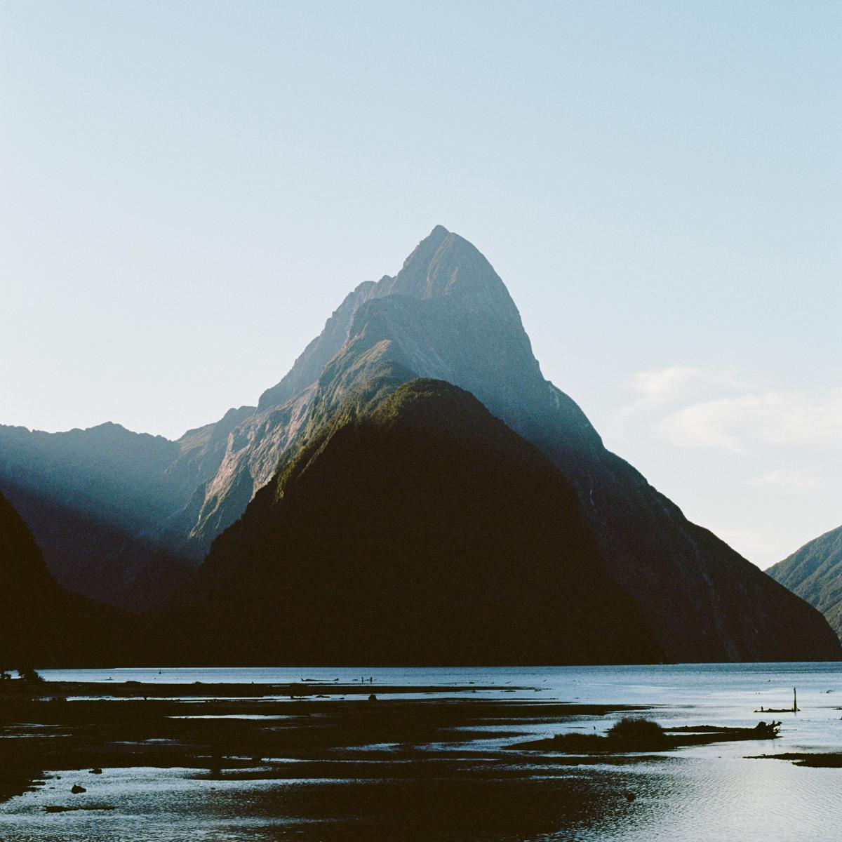 A large mountain peak rises above a body of water with a rocky shore.