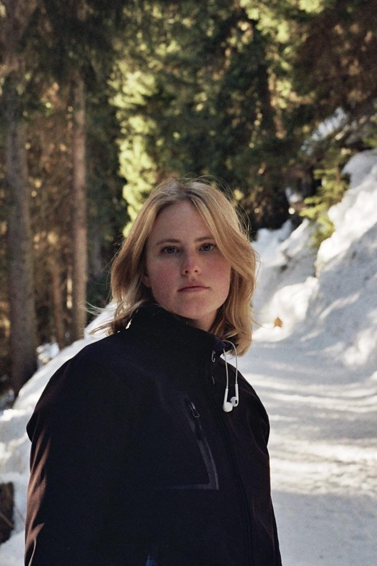 A woman with blonde hair and a black jacket stands on a snowy path in a forest.