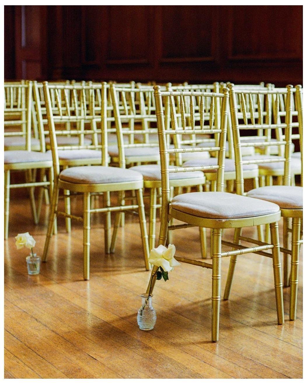Rows of golden chairs with white roses in small vases are arranged on a wooden floor.