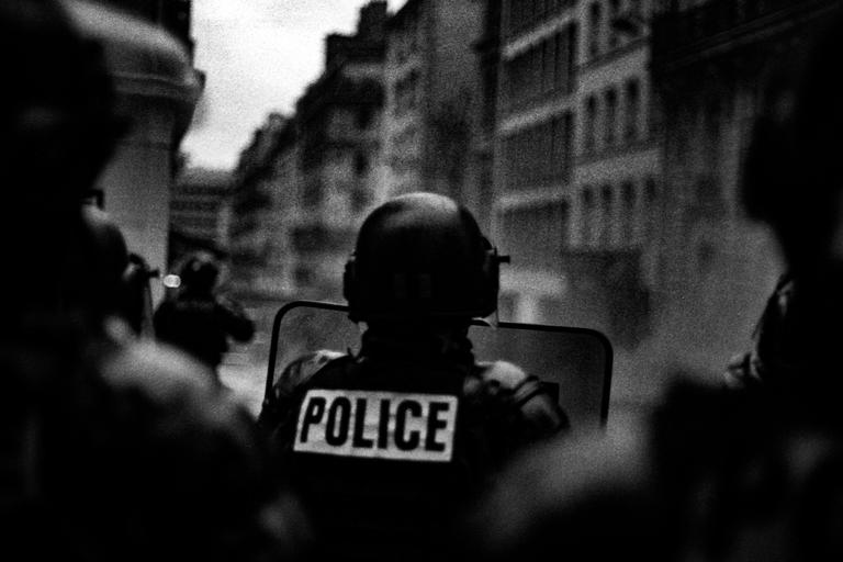 A police officer in riot gear with a shield stands in the street with buildings in the background.