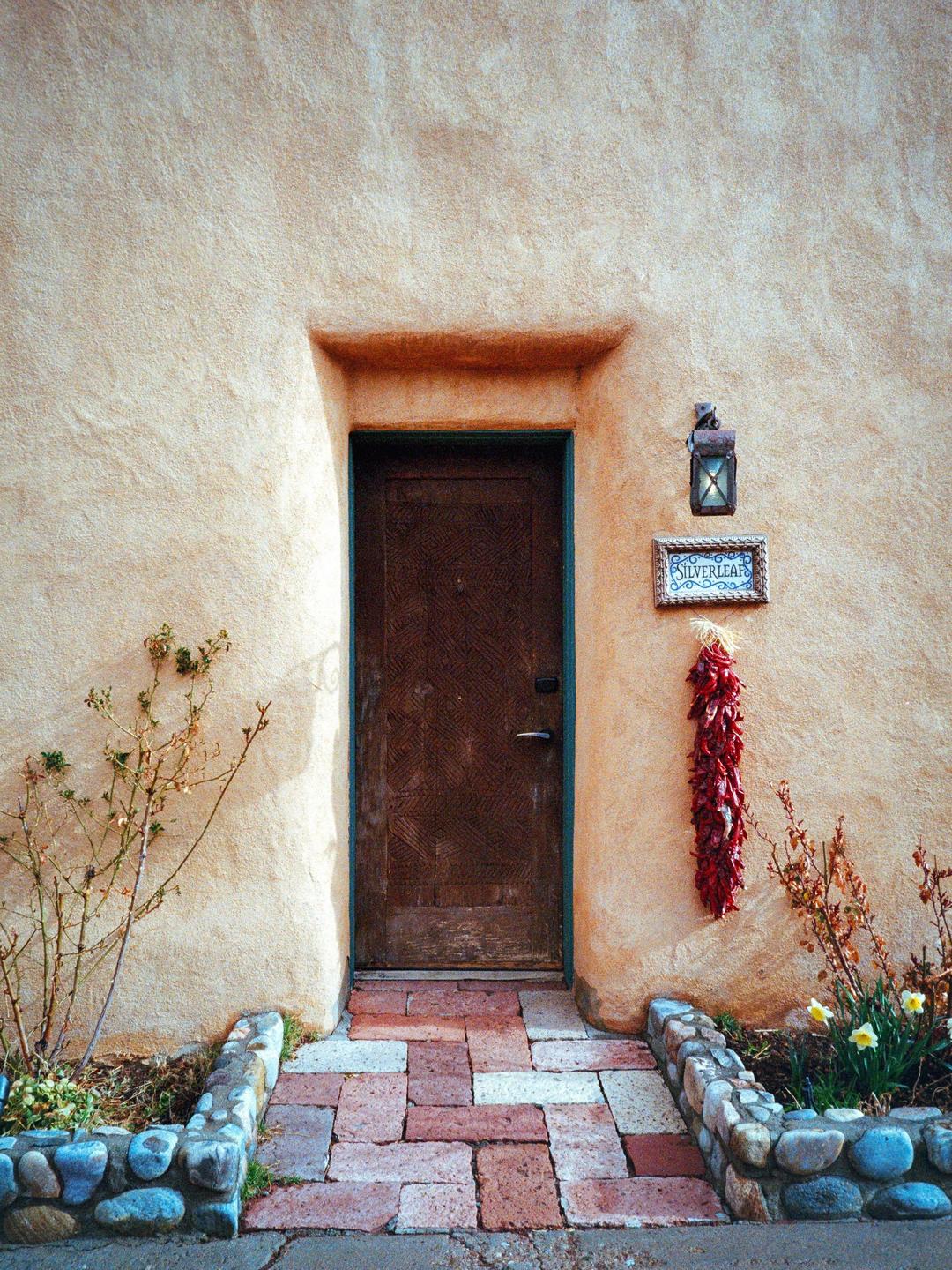 A wooden door is set into a stucco wall with a brick path leading to it, flanked by small stone walls and plants.