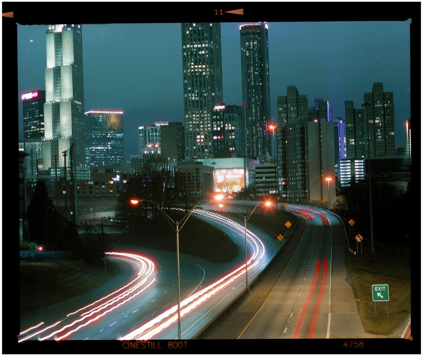 A long exposure captures light trails from vehicles on a highway at night, with a city skyline in the background.