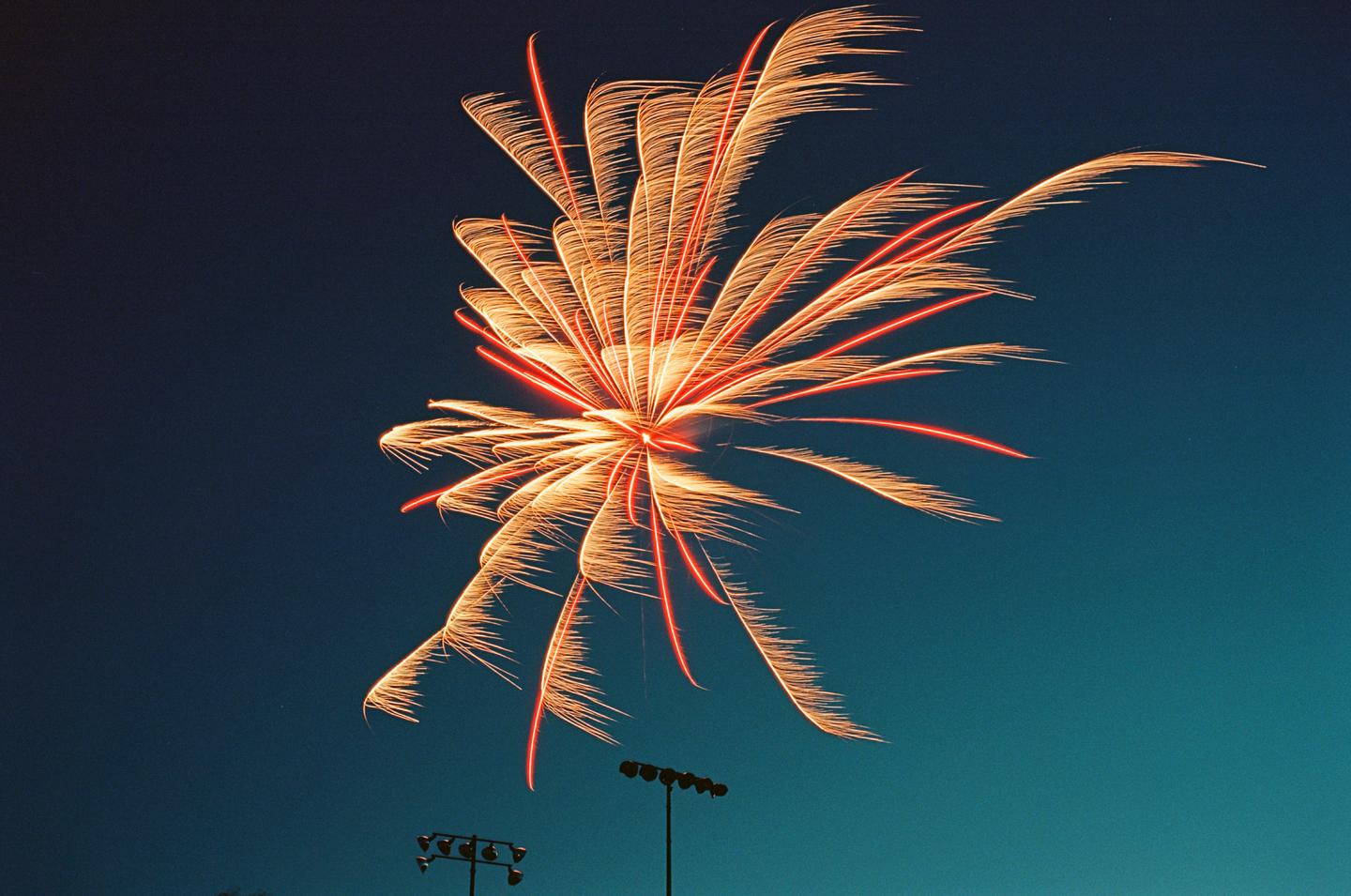 A bright orange and red firework explodes in the night sky above stadium lights.