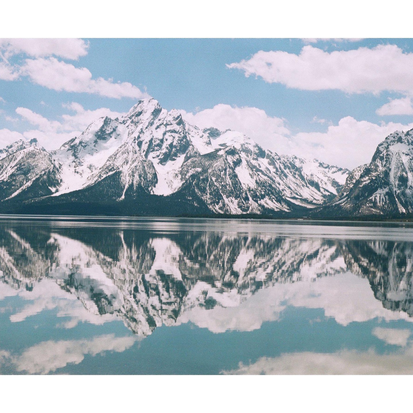 Snow-capped mountains are reflected in a calm lake under a blue sky with clouds.