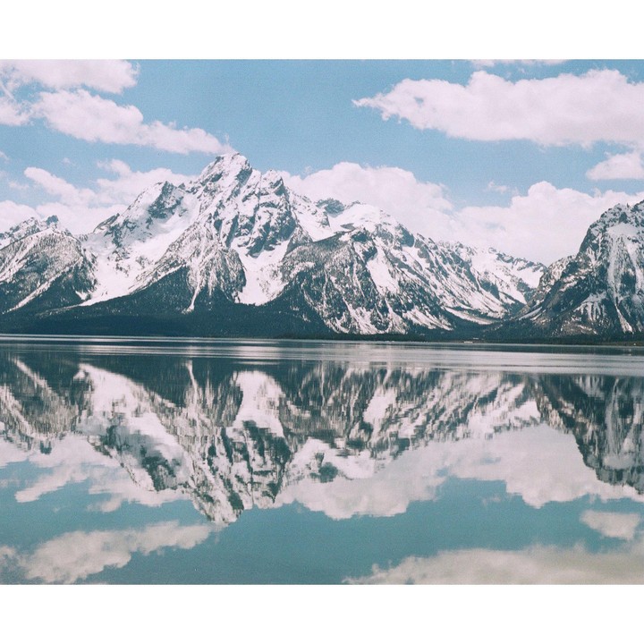 Snow-capped mountains are reflected in a calm lake under a blue sky with clouds.
