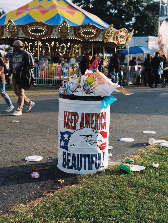 An overflowing trash can with "Keep America Beautiful" written on it stands in front of a carousel at what appears to be a fairground.