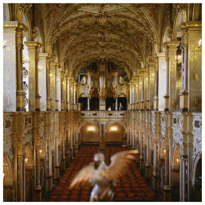A grand hall with ornate golden decorations, an organ, and rows of empty seats is viewed from above, with a blurred statue in the foreground.