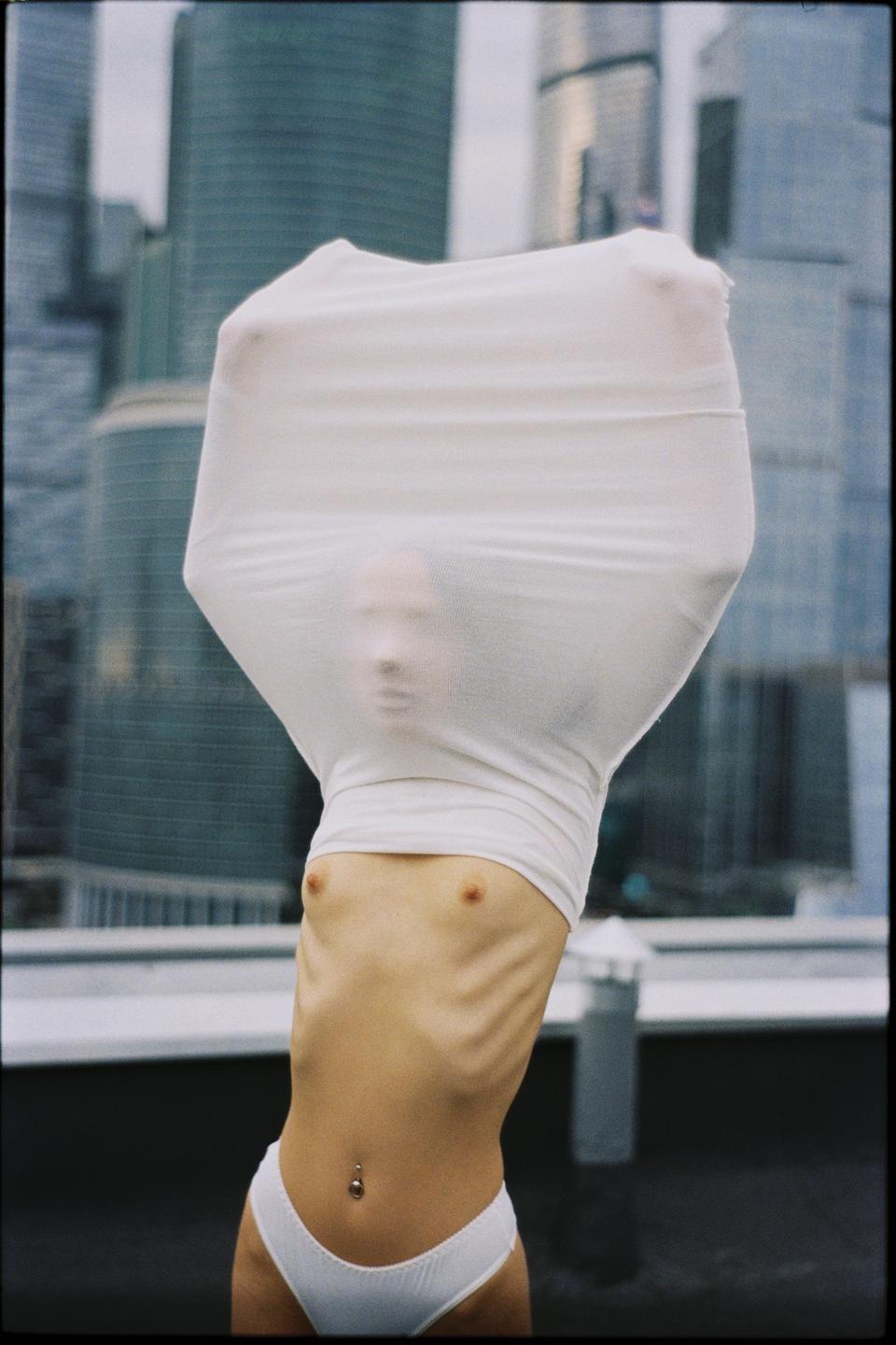 A woman with her head covered by a white fabric stands in front of tall buildings.