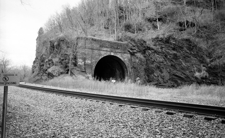 A black and white photograph shows a brick train tunnel entrance next to a railway track, with a "No Trespassing" sign in the foreground.