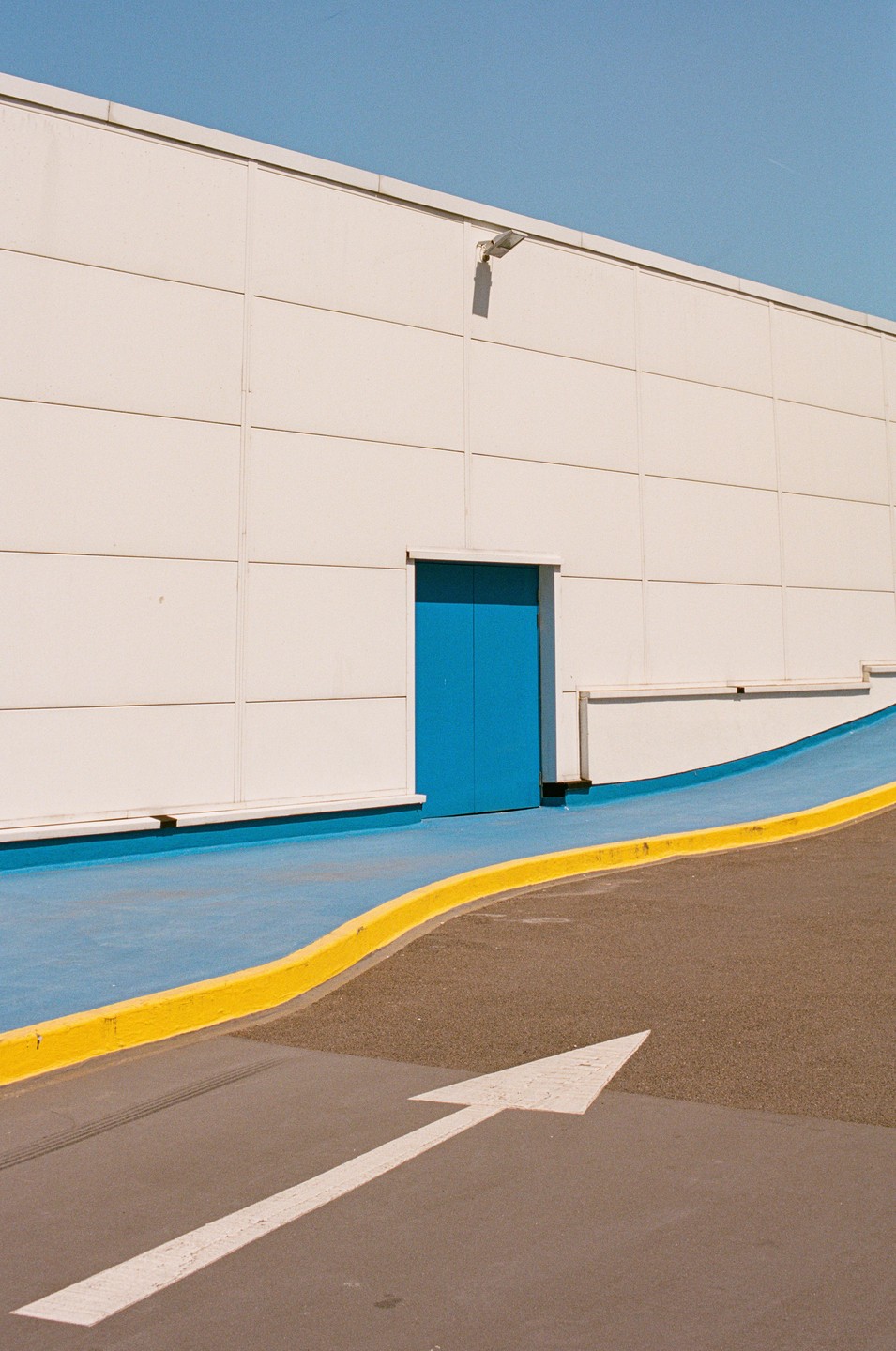 A white building with a blue door and a blue ramp with a yellow curb is shown under a clear sky.