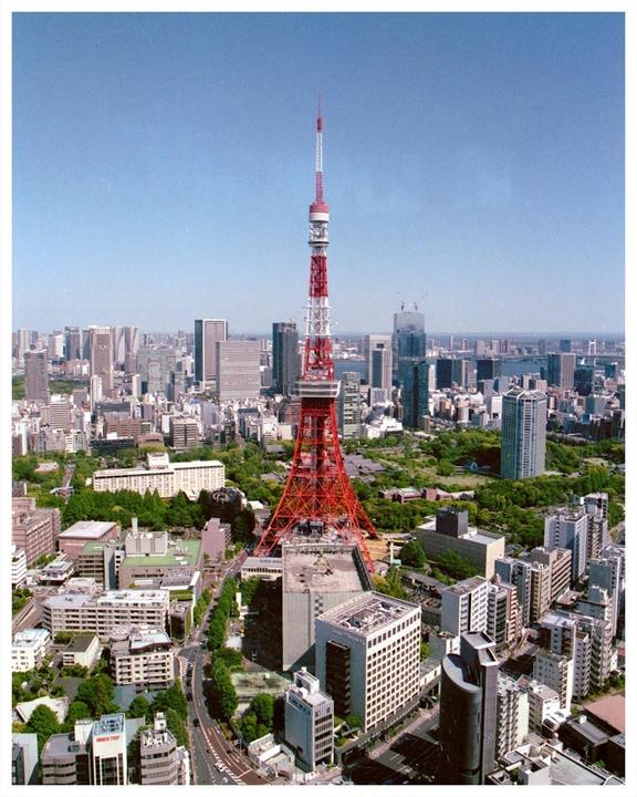 An aerial view shows the Tokyo Tower surrounded by a dense urban landscape with numerous buildings and green spaces under a clear sky.