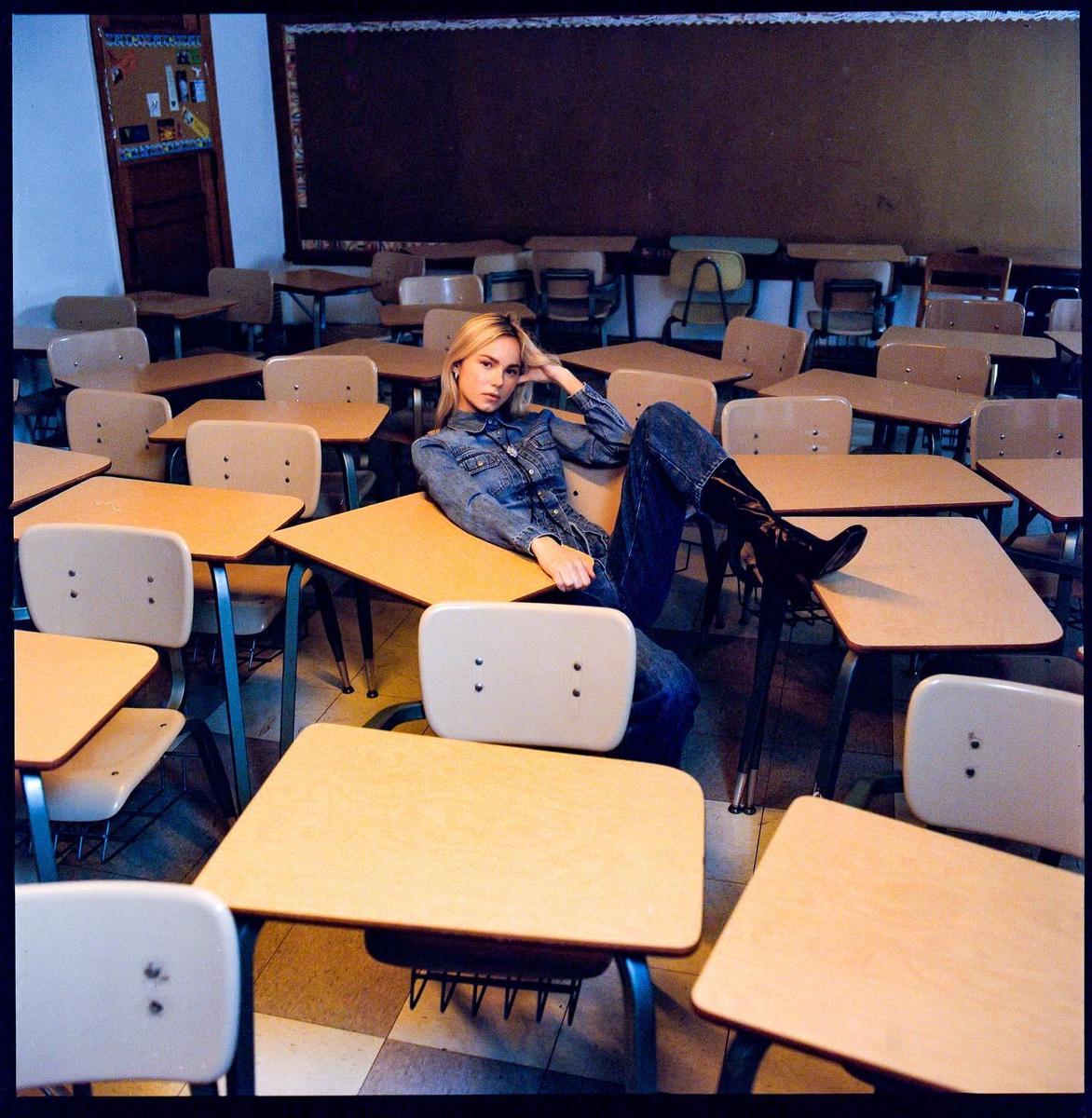 A woman in a denim jumpsuit and boots reclines on a desk in a classroom.