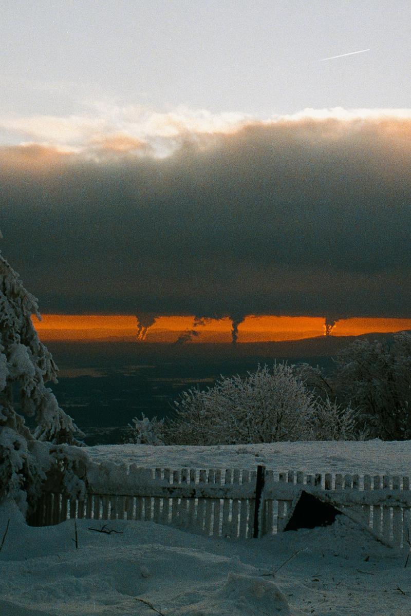A snow-covered landscape with a wooden fence in the foreground and a bright orange sunset visible through a break in the clouds.