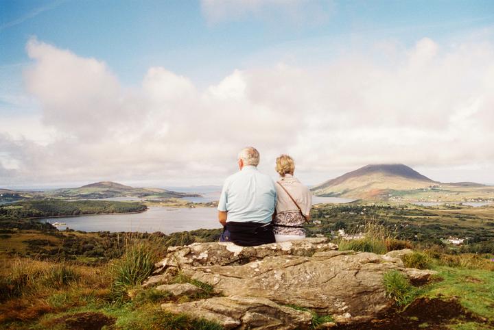 An elderly couple sits on a rock overlooking a body of water and mountains under a cloudy sky.