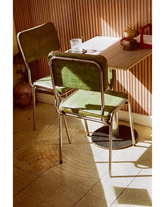 Two green chairs are pulled up to a small table with a glass on it, illuminated by sunlight streaming through a window.