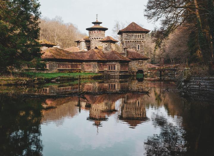 A stone building with a red tile roof is reflected in the still water of a lake, surrounded by trees.