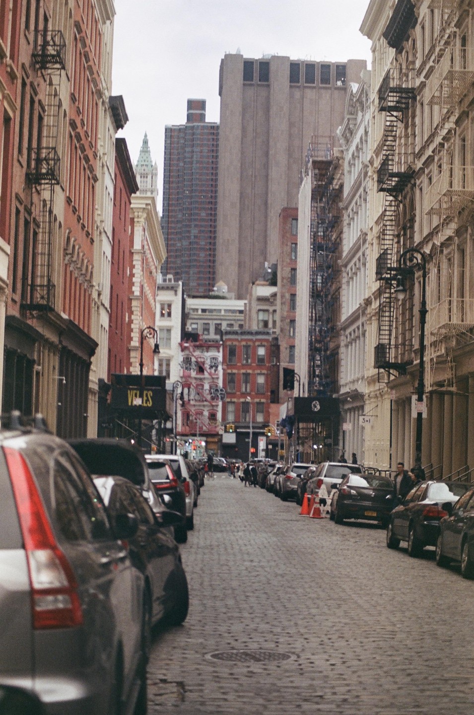 A narrow cobblestone street is lined with parked cars and tall buildings under an overcast sky.