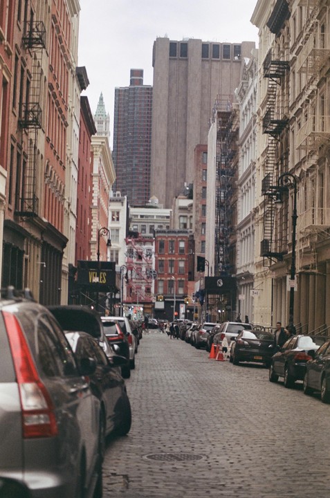 A narrow cobblestone street is lined with parked cars and tall buildings under an overcast sky.