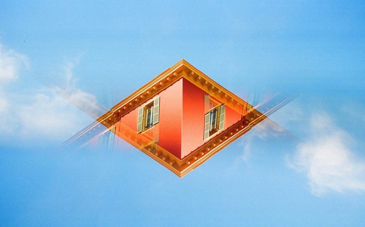 A double exposure shows a building corner with windows and shutters against a blue sky with clouds.