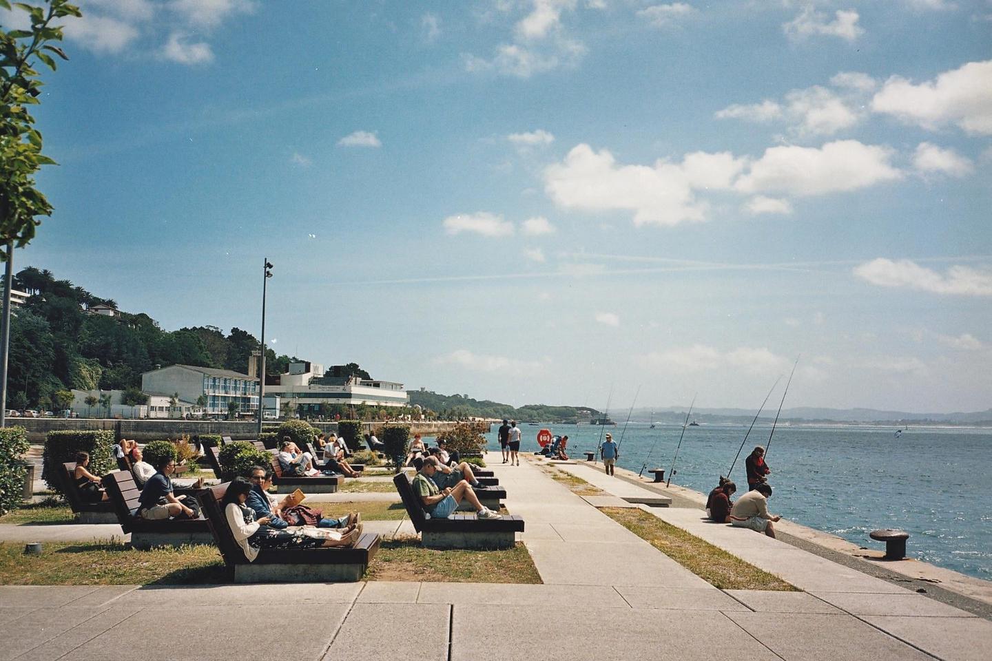 People relax on benches and fish along a waterfront promenade with buildings and hills in the background.