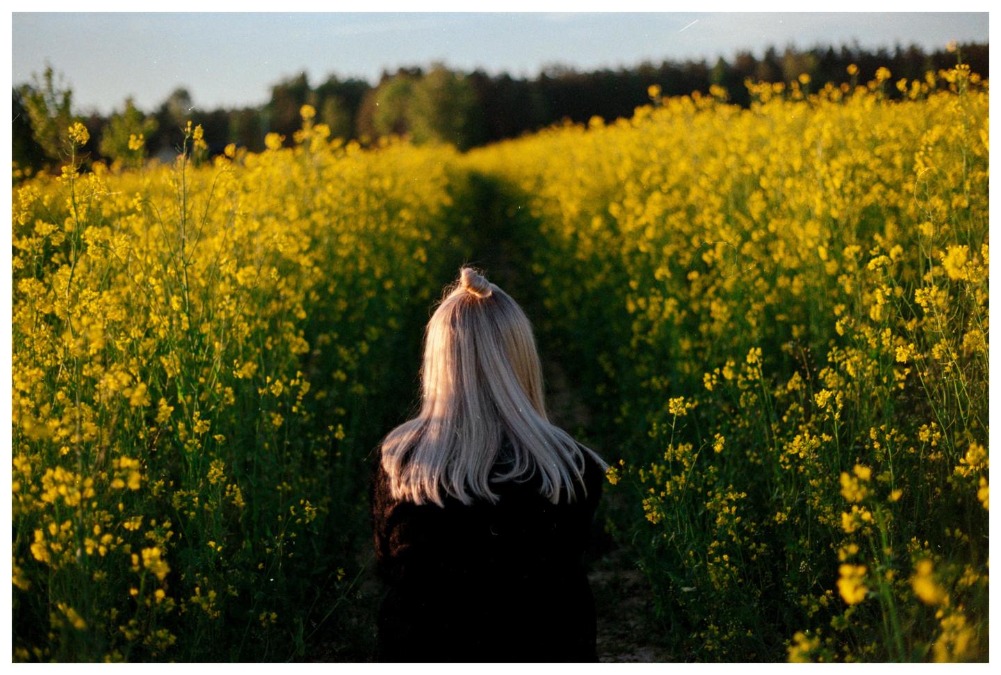 A woman with long blonde hair stands in a field of yellow flowers, looking away from the camera.