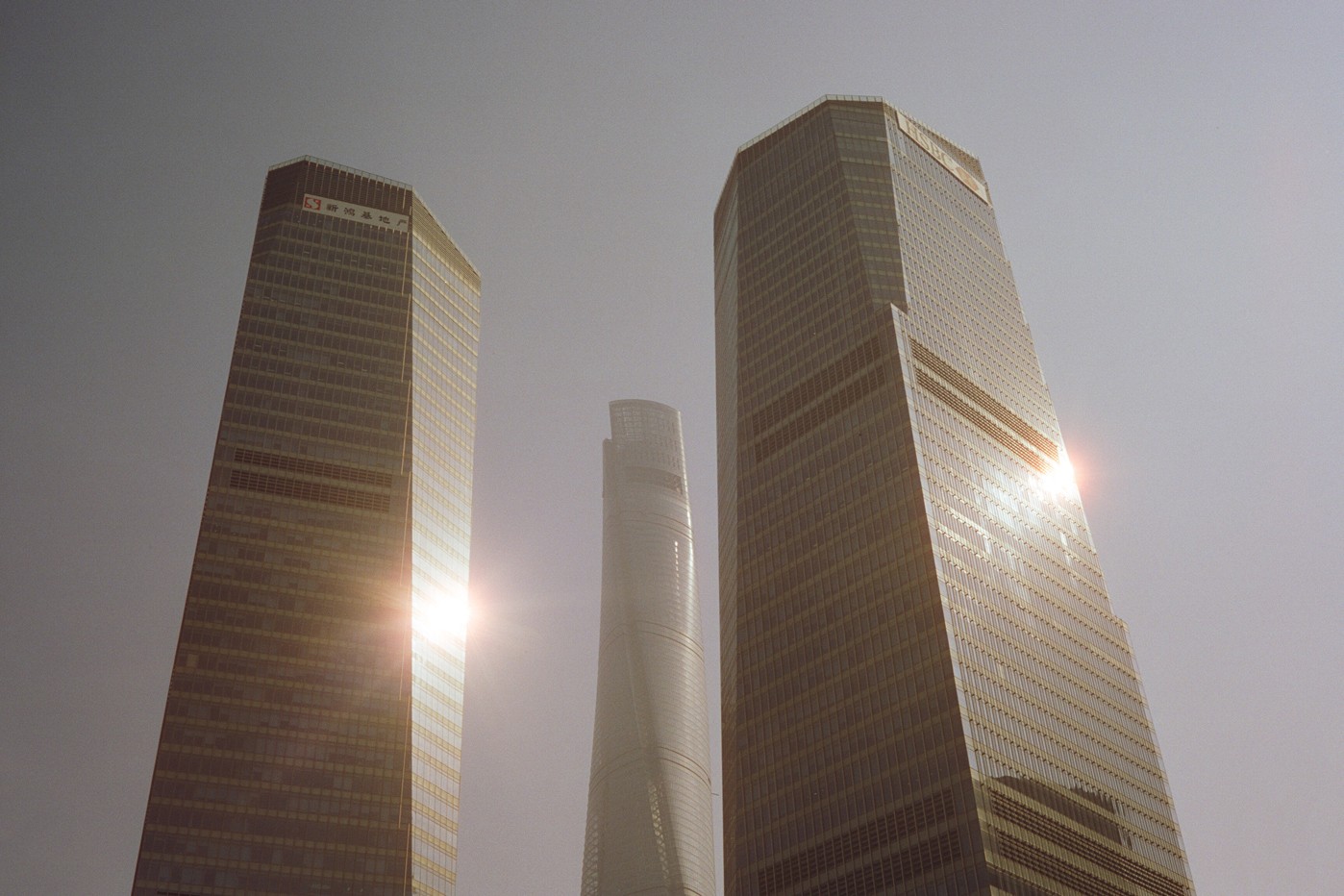 Three skyscrapers are visible from a low angle, with two on either side of a taller, more slender building in the center, and sun flares reflecting off the glass facades.