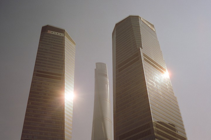 Three skyscrapers are visible from a low angle, with two on either side of a taller, more slender building in the center, and sun flares reflecting off the glass facades.