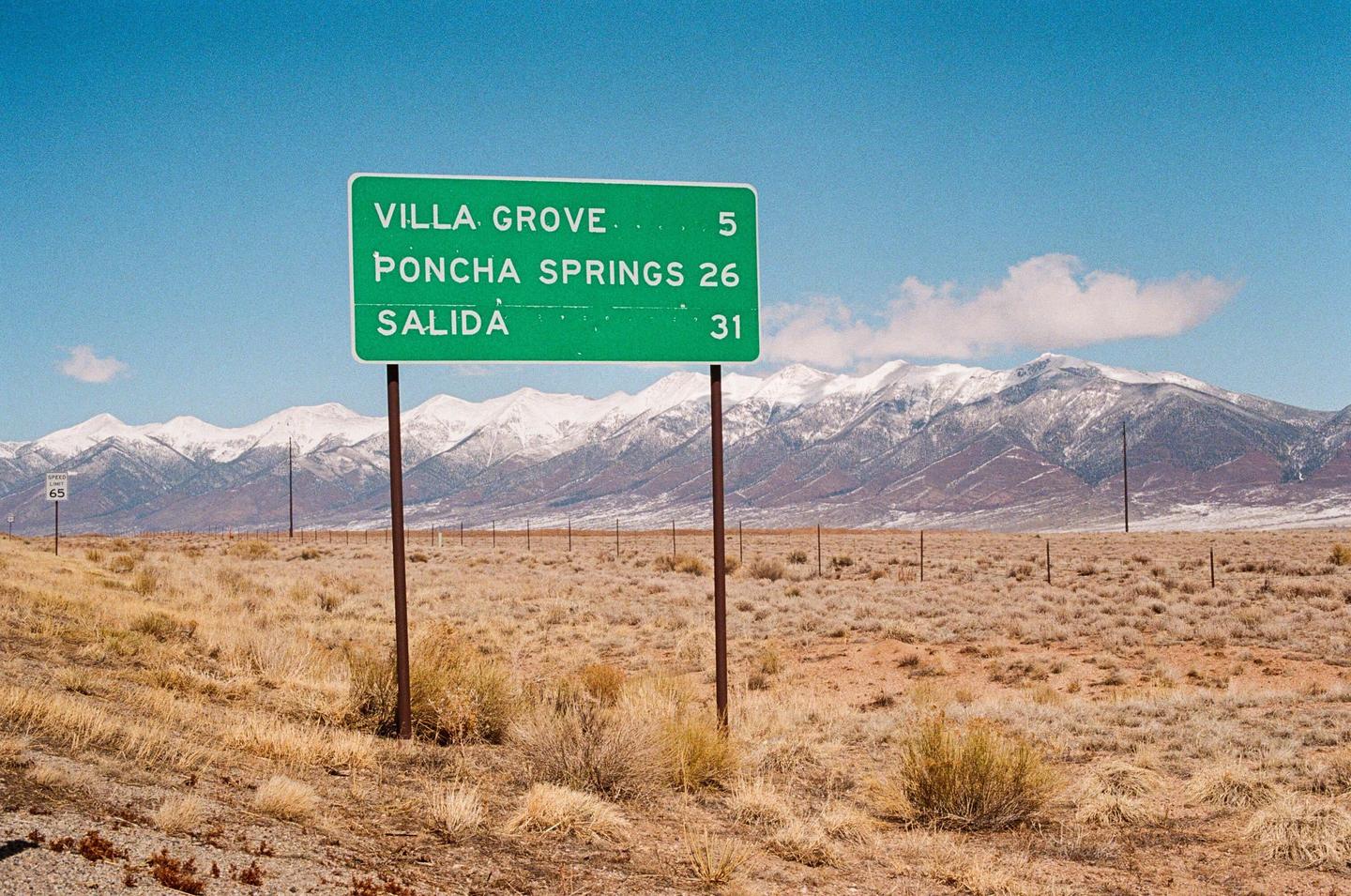 A green road sign stands in a dry, grassy field with snow-capped mountains in the background under a clear blue sky.