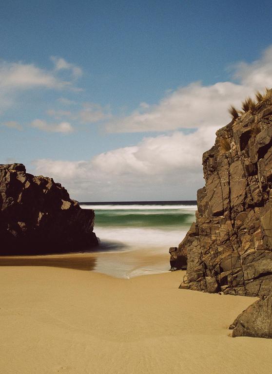 A sandy beach is framed by two rocky cliffs with blurred waves washing ashore under a blue sky with clouds.