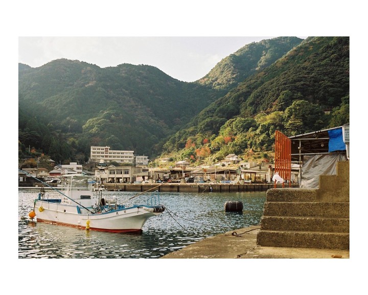 A white fishing boat is docked in a harbor with a town and lush green mountains in the background.