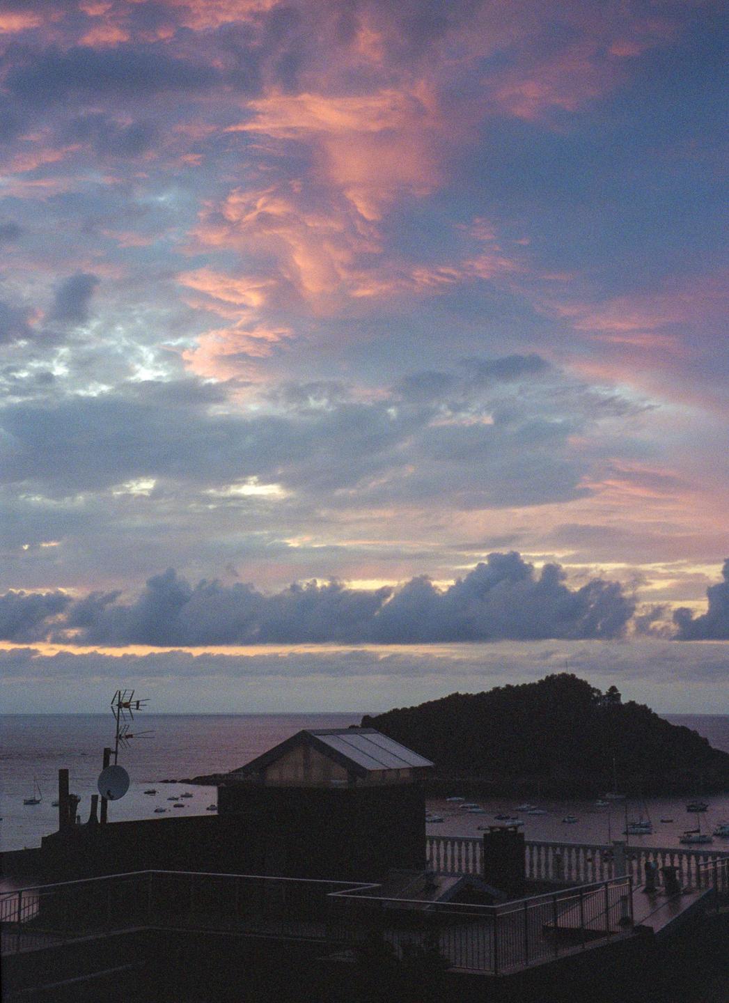 A dramatic sunset with pink and purple clouds hangs over a bay with boats and a dark island.
