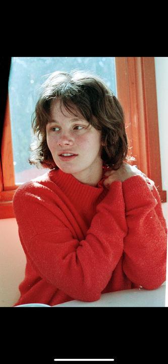 A woman in a red sweater looks out a window, with light shining on her hair.