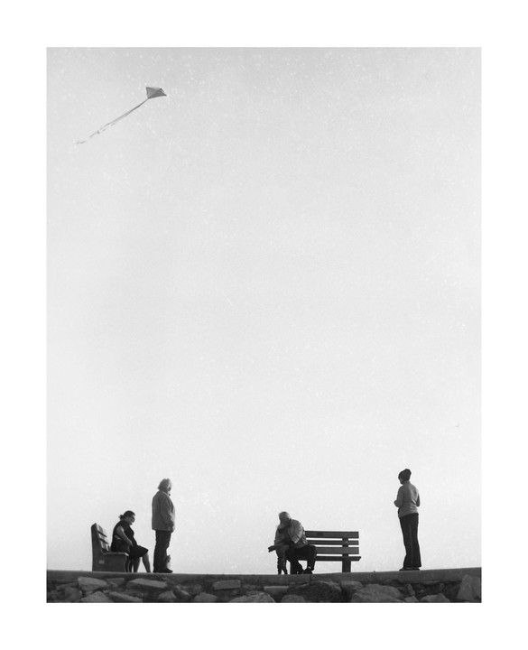 Four people are gathered on a stone wall with a kite flying high in the sky.