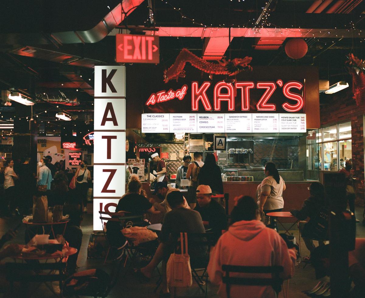 People are seated at tables and standing in line at a food hall with a large red neon sign that says "Katz's".