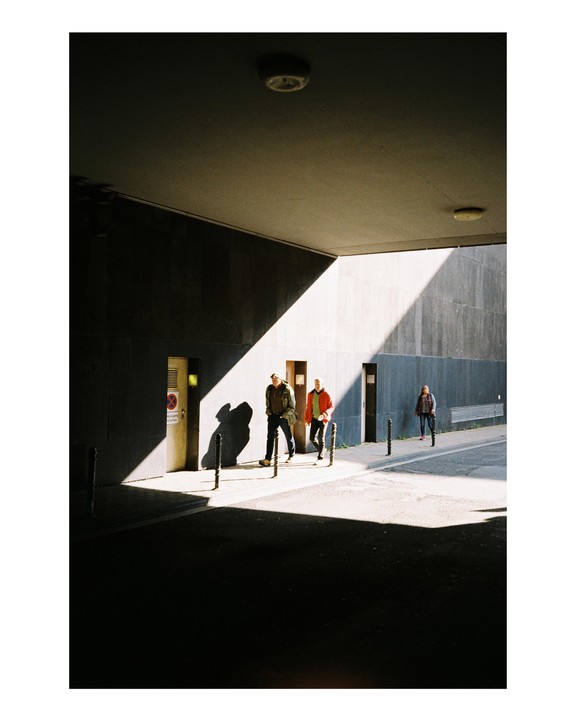 Two men walk through a sunlit passage while another person stands in the shadow.