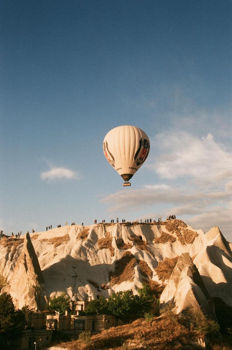 A hot air balloon floats above a rocky landscape with buildings and people on a hill.